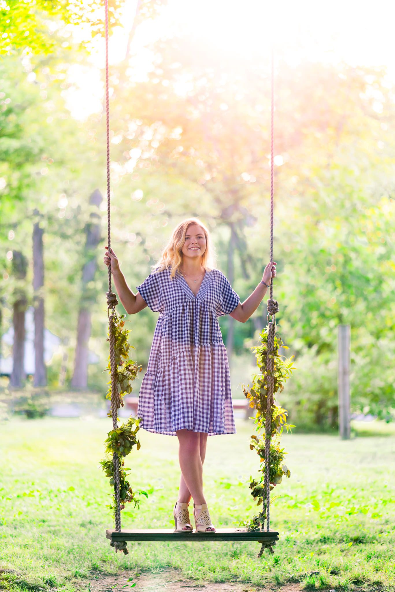 senior picture of a young woman in blue and white dress standing on a swing with floral ropes at sunset