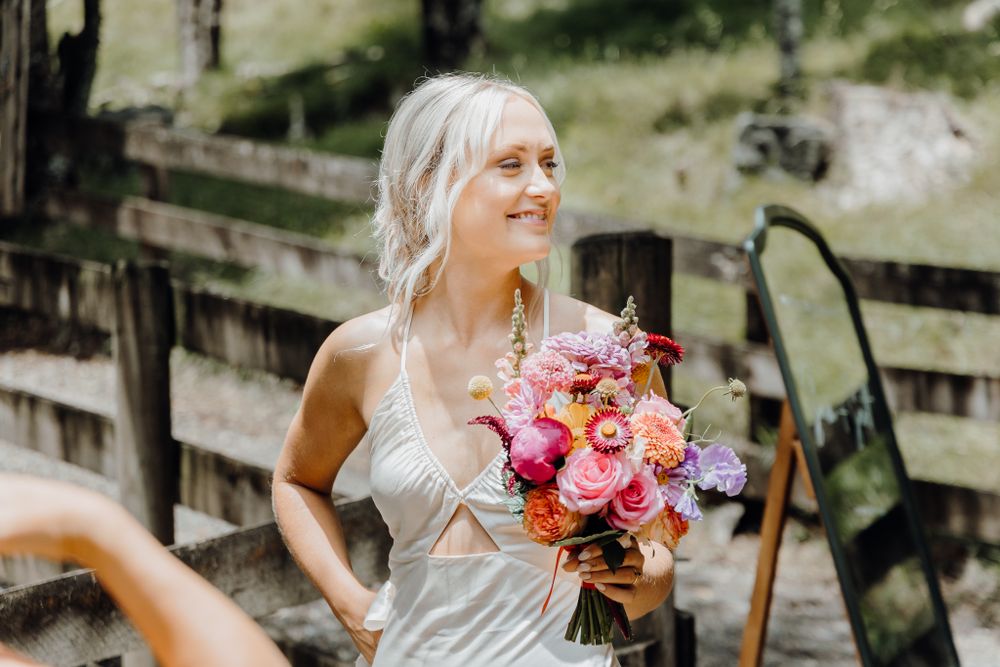 Bride holds vibrant pink and coral bouquet while smiling during outdoor wedding.