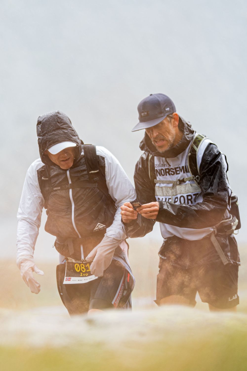 Two runners in vests brave stormy conditions on a misty trail during an endurance race.