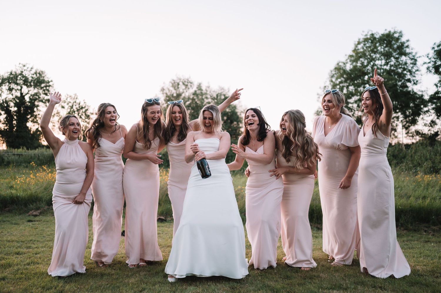 A wedding party in pale pink bridesmaid dresses and white bridal gown pose together outdoors at sunset.