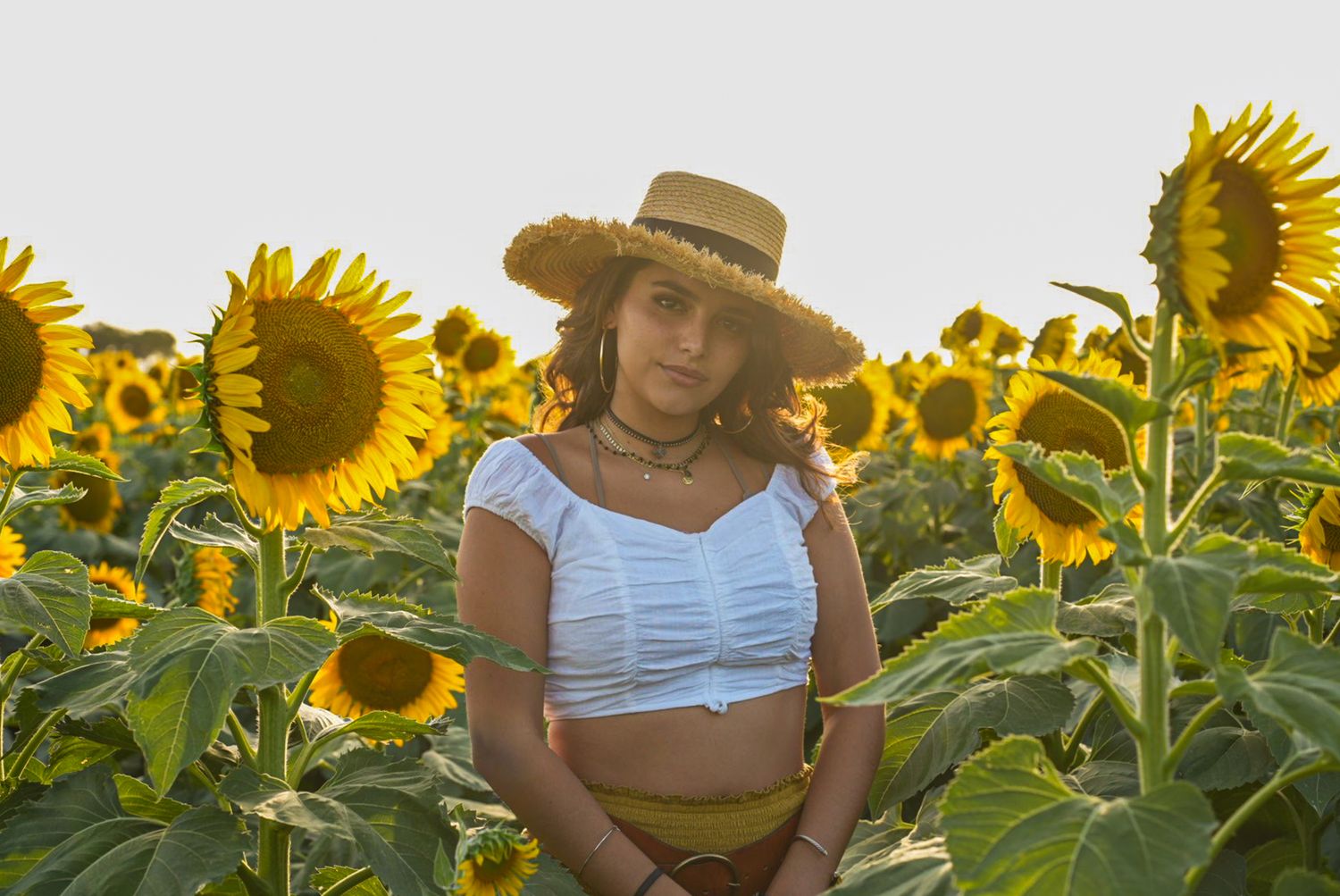 Person in summer attire poses thoughtfully in a vibrant sunflower field at golden hour.