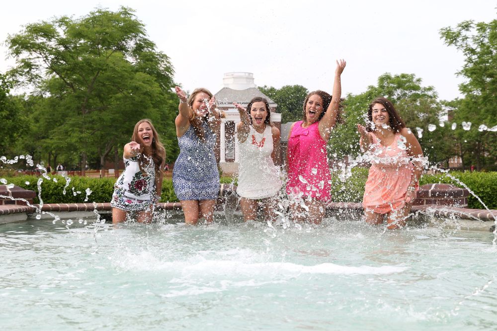 Group of friends splash and jump in fountain during graduation celebration.