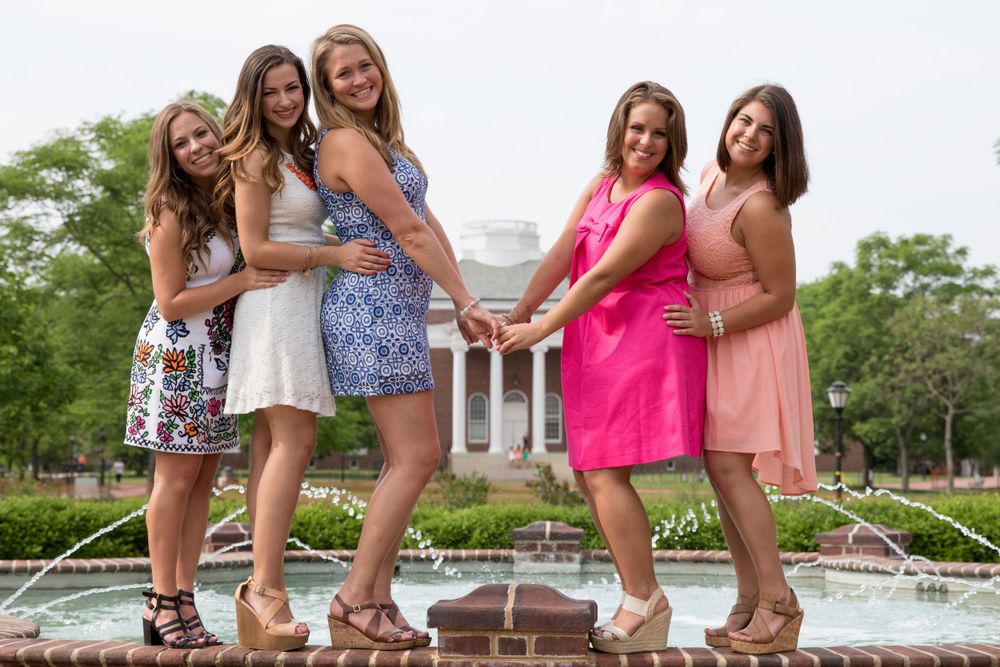 Friends in summer dresses pose playfully by campus fountain with classic building in background.
