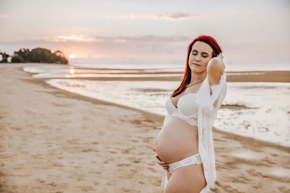 beautiful pregnant pregnant lady with bright red hair on the beach showing off her bump at the beach for a photoshoot