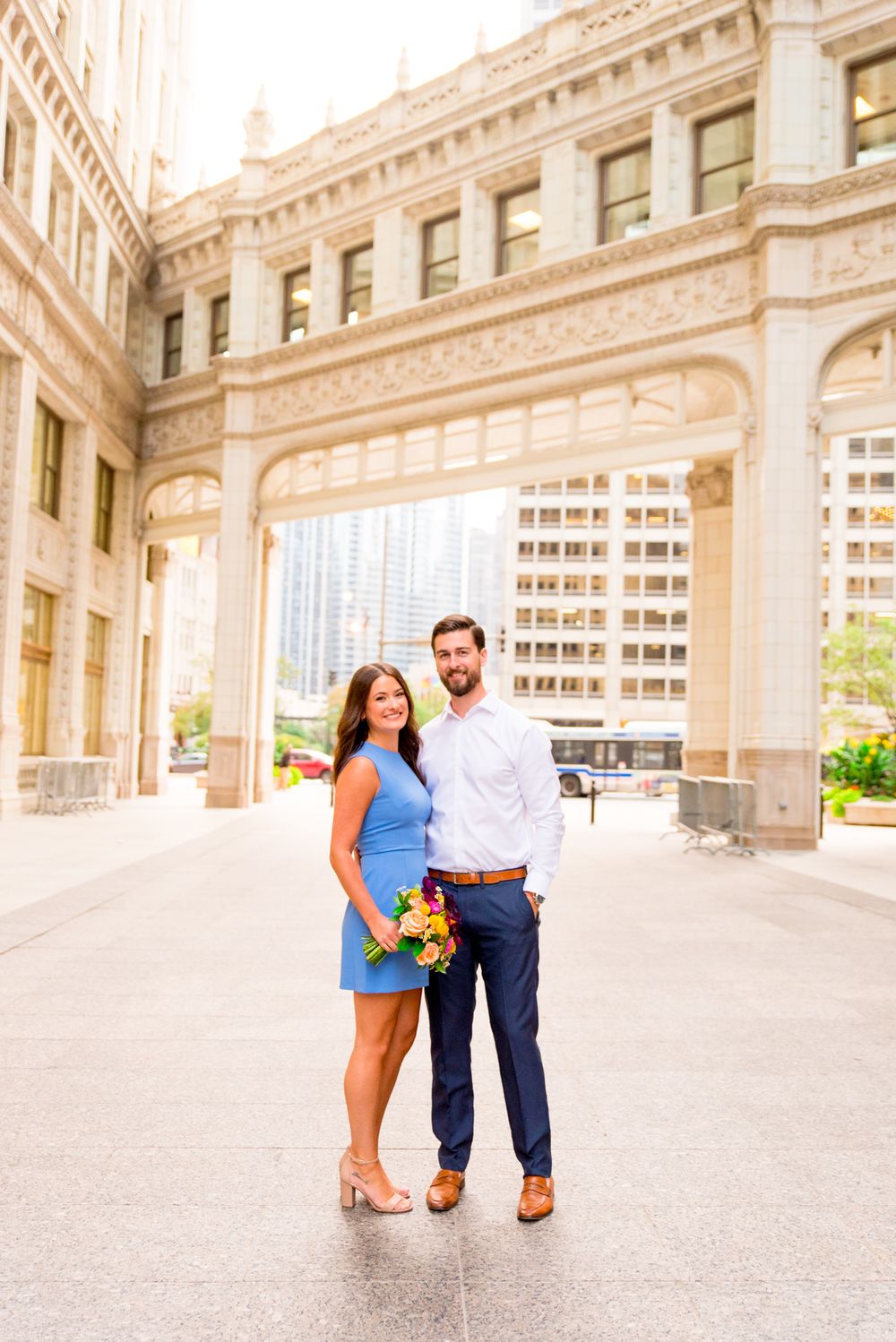 woman with brown hair in a light blue dress holding hands with fiance in front of the wrigley building in chicago