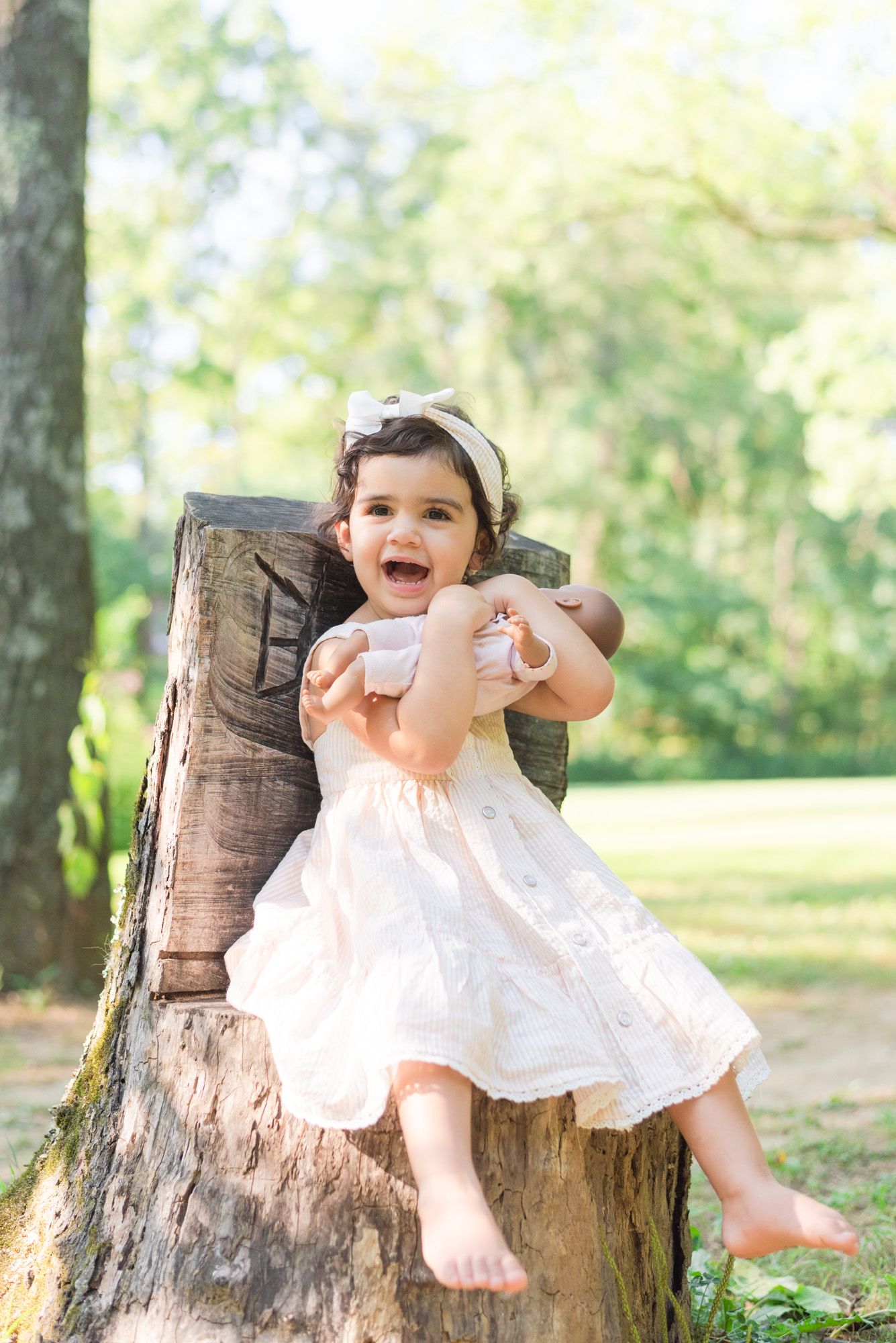 Toddler laughing sitting on stump holding her baby doll at Emmerling Park in Cheswick Pennsylvania