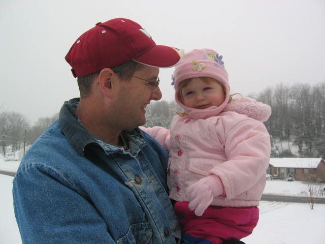 Winter scene with adult holding young child bundled in pink snowsuit.