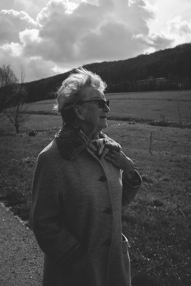 A person in sunglasses and coat gazes upward on a rural path with hills in the background.