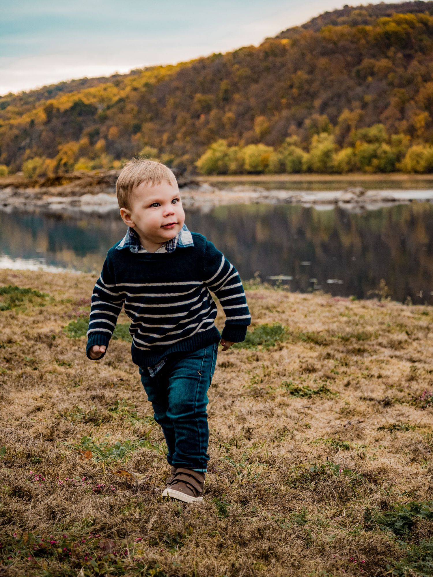 Child in striped sweater exploring near an autumn lake with mountains in the background.