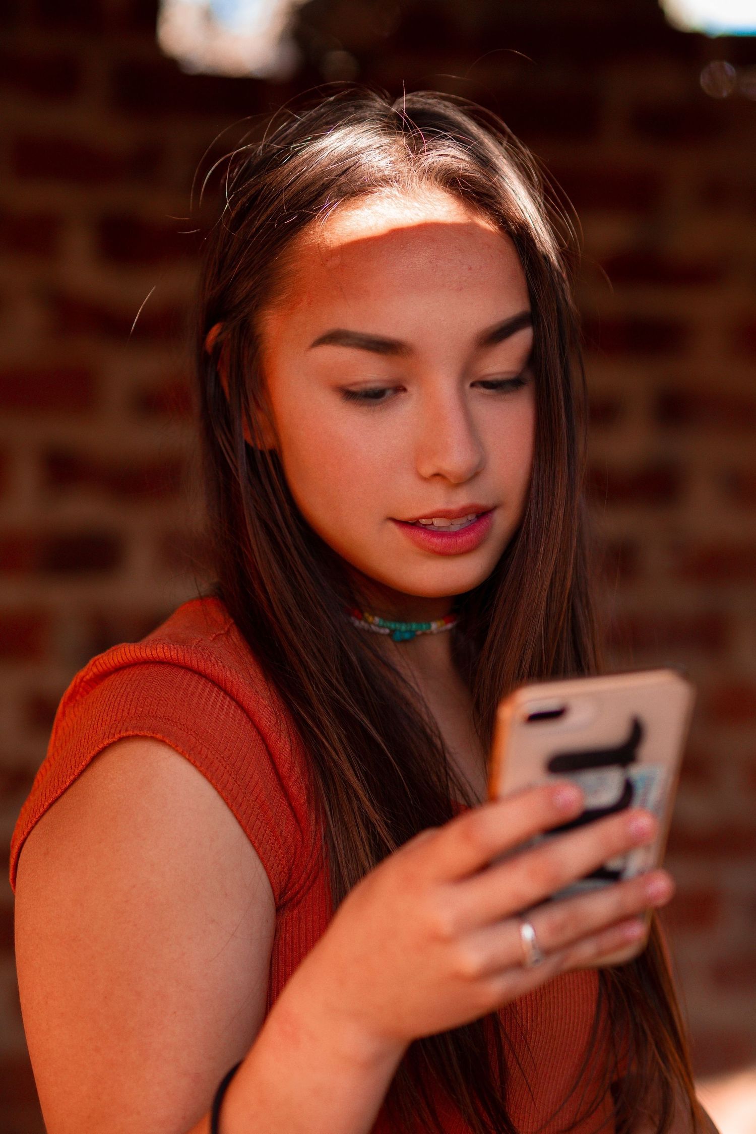 Woman in red top looking down at smartphone against brick wall background.