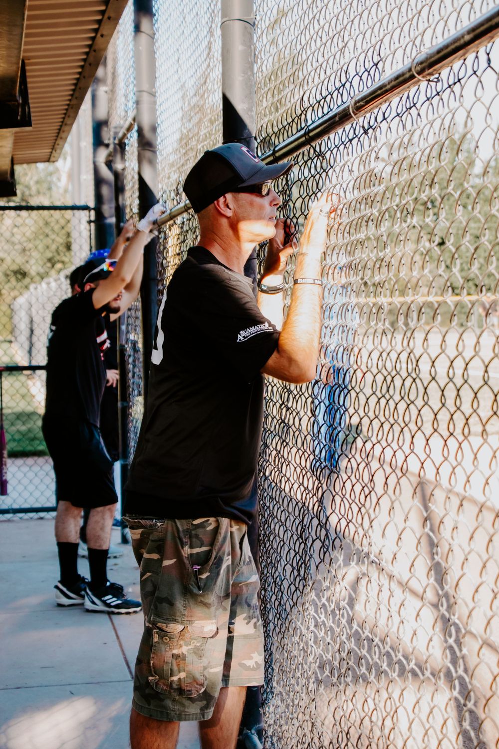 Baseball player in black shirt and camo shorts standing by chain link fence during practice.