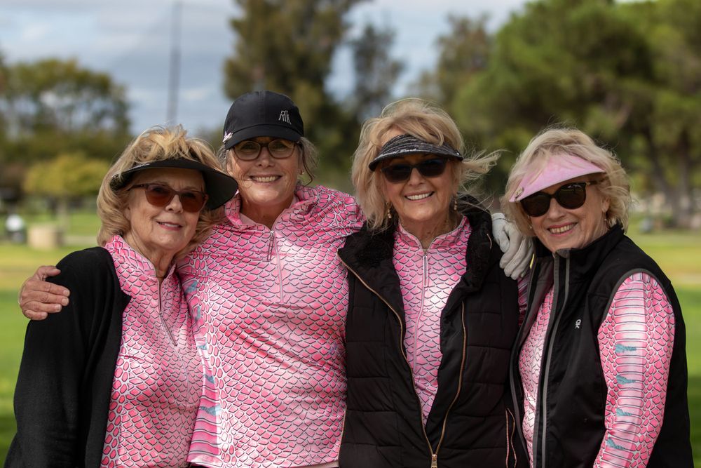 Four golfers wearing pink and black athletic gear smiling together on a sunny day at the golf course.