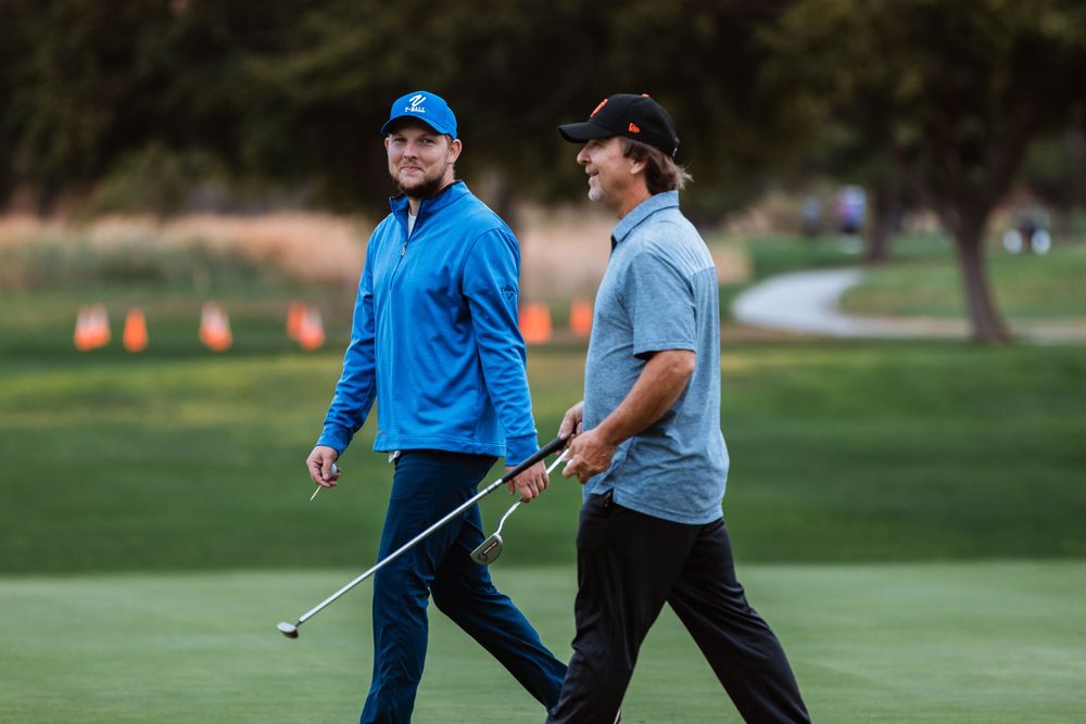 Two golfers walk across a lush green course carrying clubs.