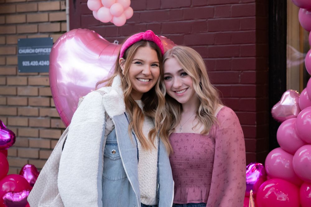 Two people smile together in front of a pink balloon display with heart shapes and festive decorations.