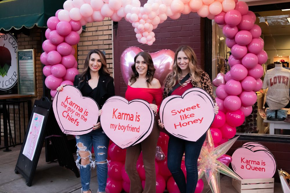 Group poses with heart-shaped signs under a pink balloon arch outside a storefront.