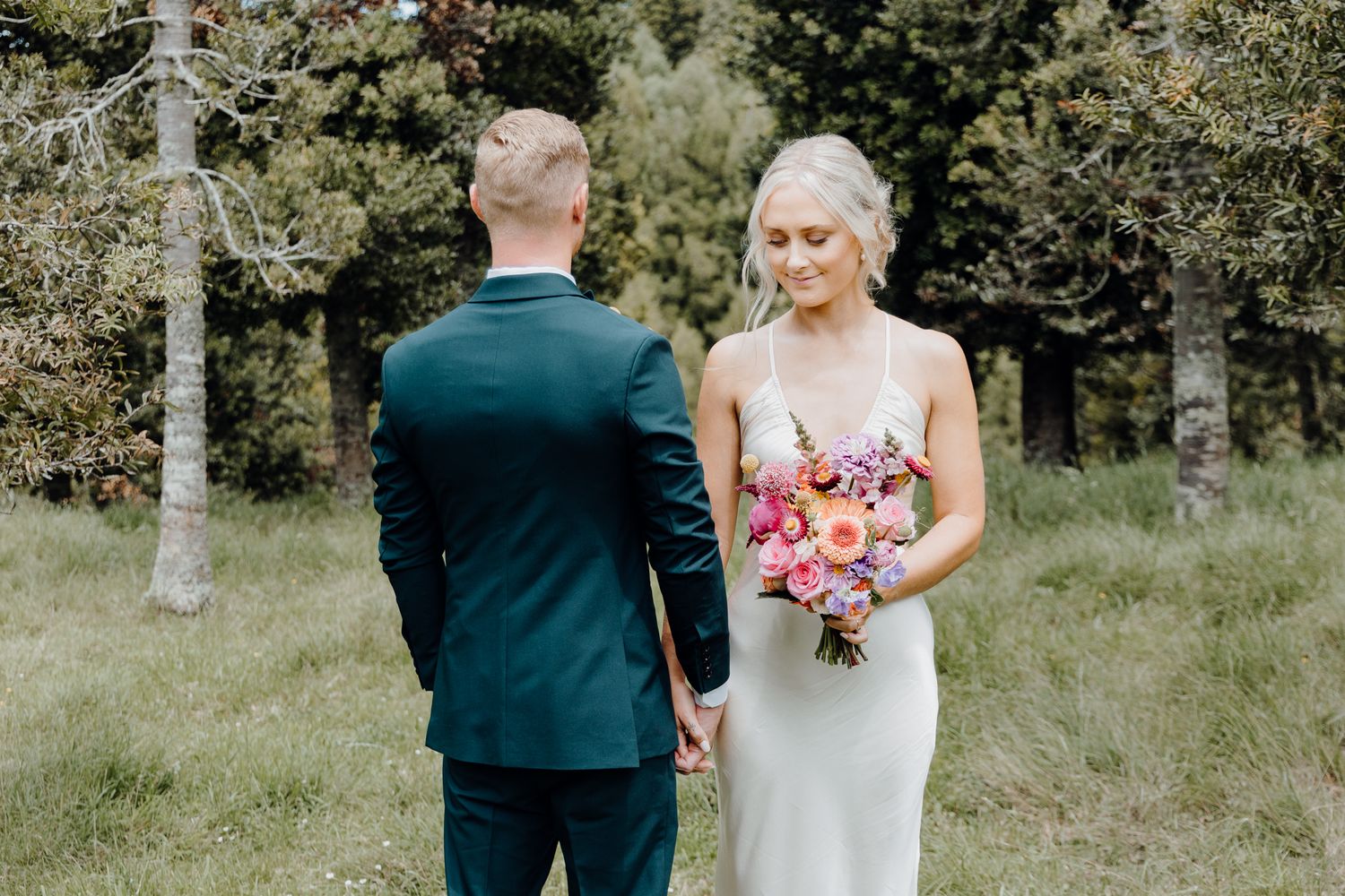 Couple shares intimate moment in outdoor wedding photos with vibrant pink bouquet and emerald suit.