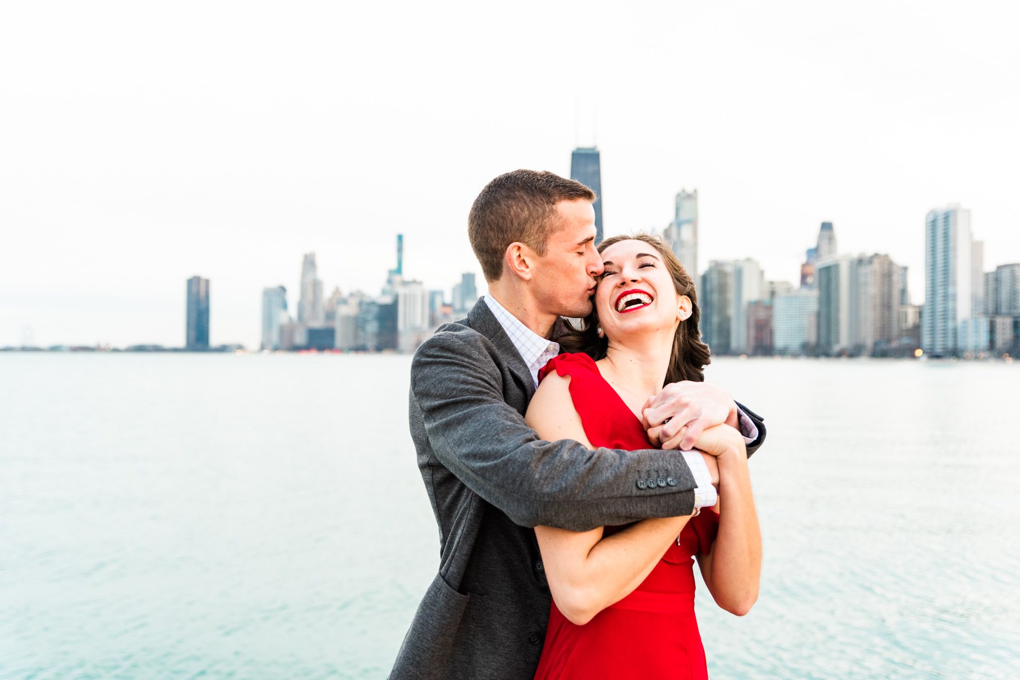 man hugging fiancee from behind and kissing her cheek in front of Chicago skyline for Chicago engagement pictures