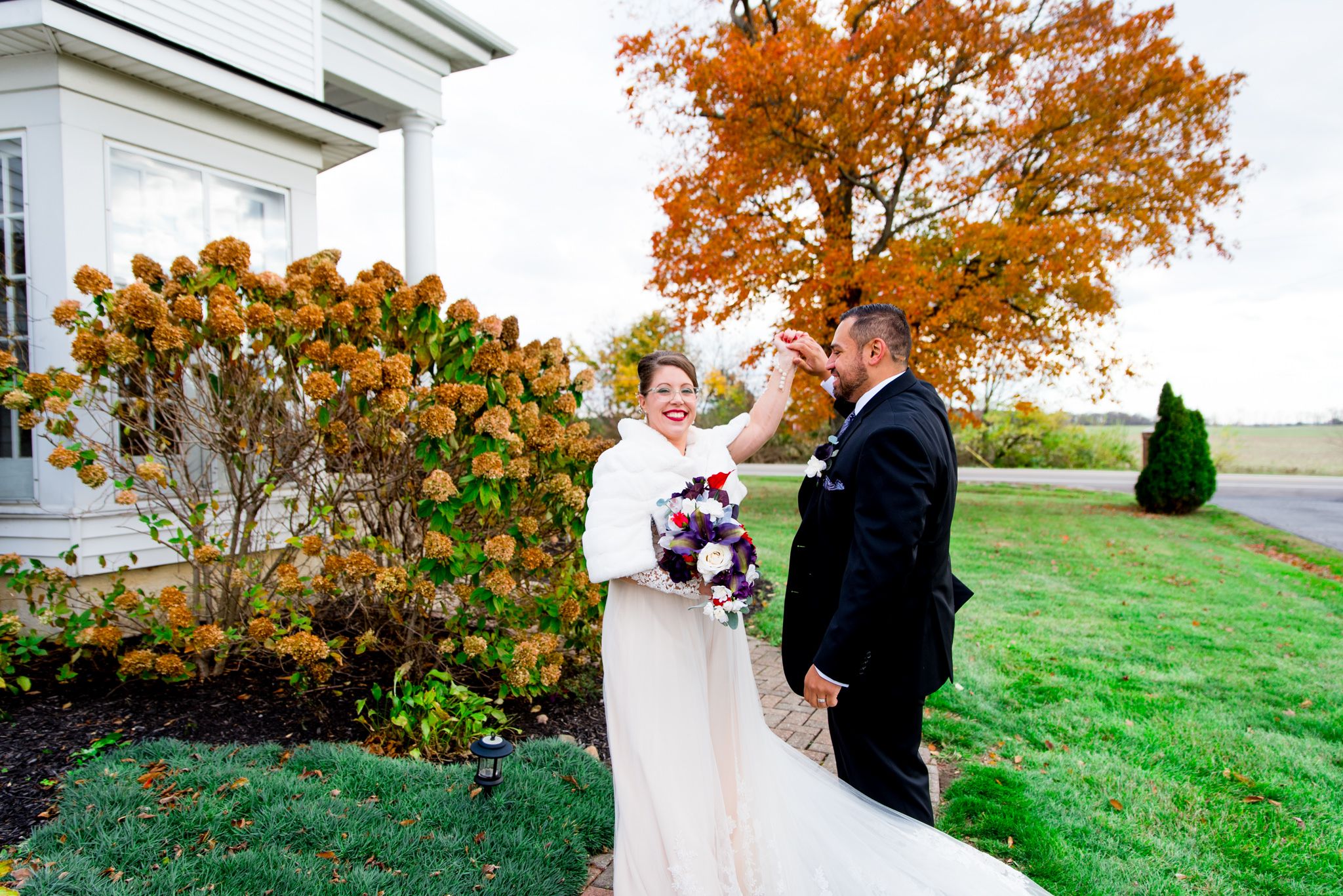 groom spinning bride around in white fur shawl and with red and purple bouquet in front of orange tree