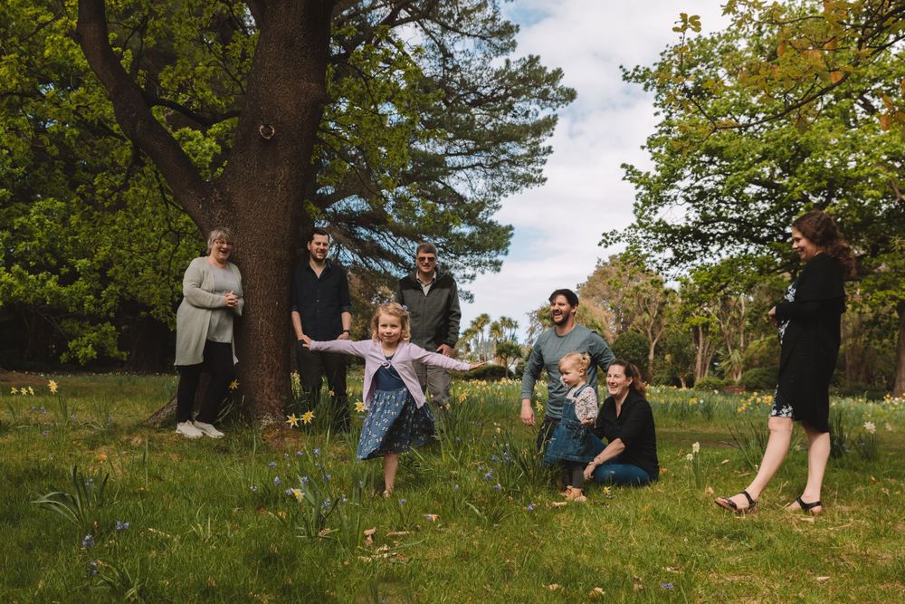 A group plays and jumps together on a grassy hill under tall trees in a park setting.