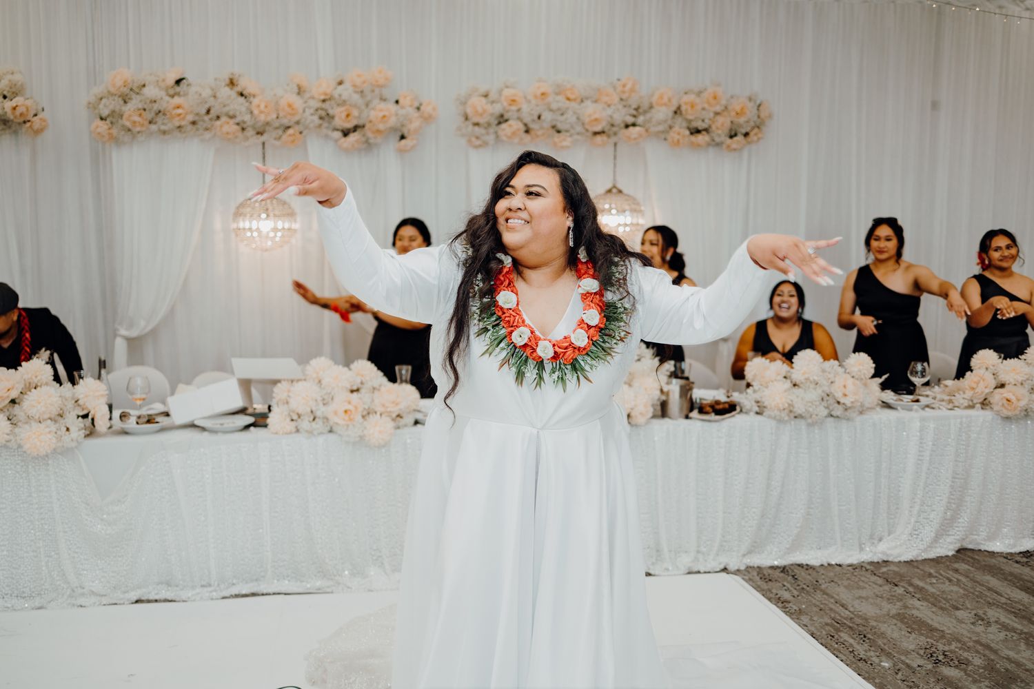 Performer in white dress with floral lei dances at wedding reception with decorative floral backdrop.