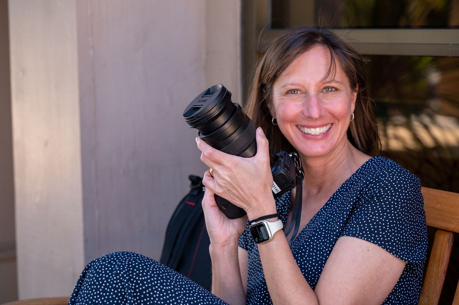 Professional photographer in a navy polka dot dress holding a camera lens with a bright smile.