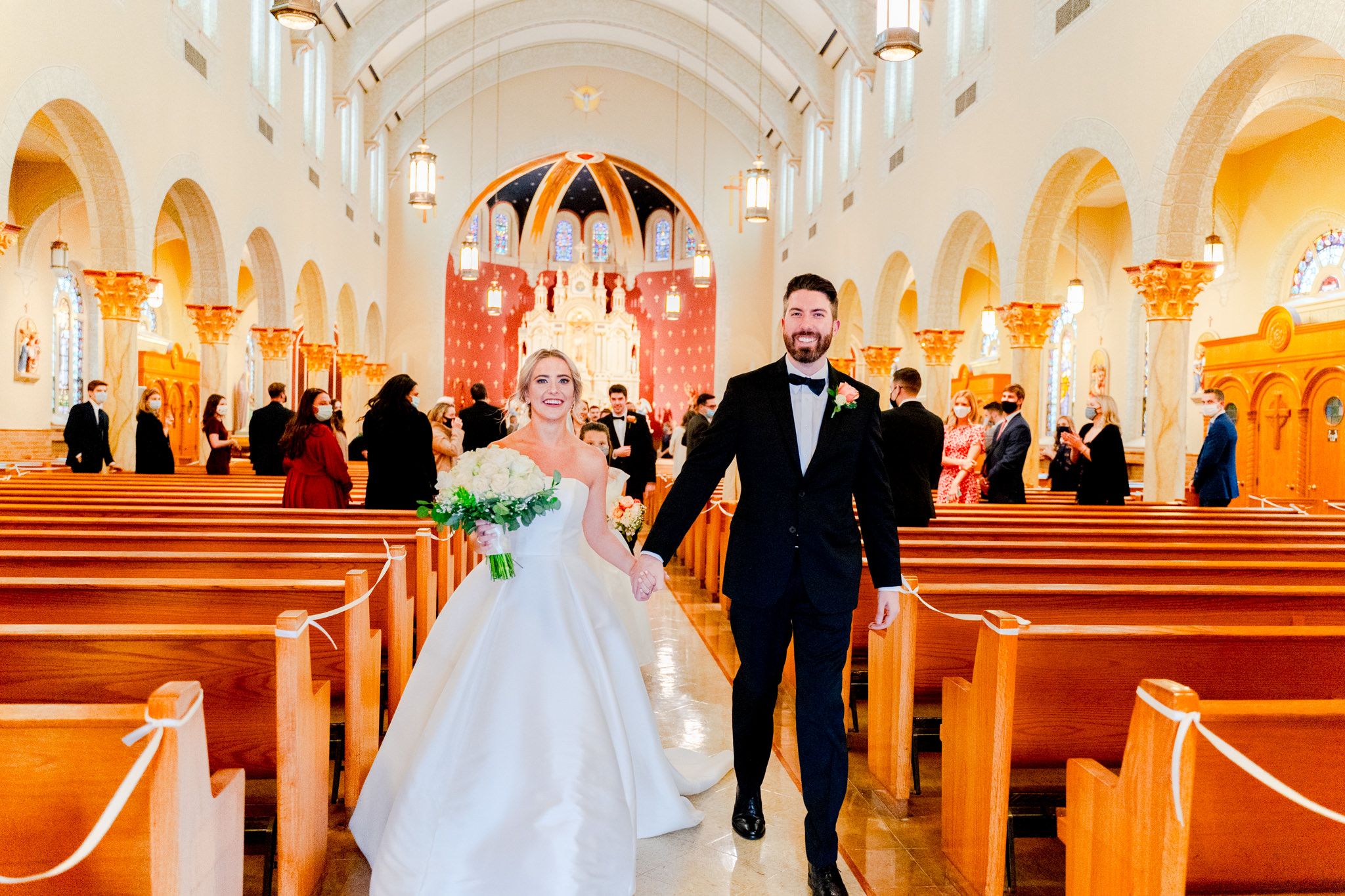 bride and groom walking down church aisle after being married, wedding guests in back smiling at them with masks on