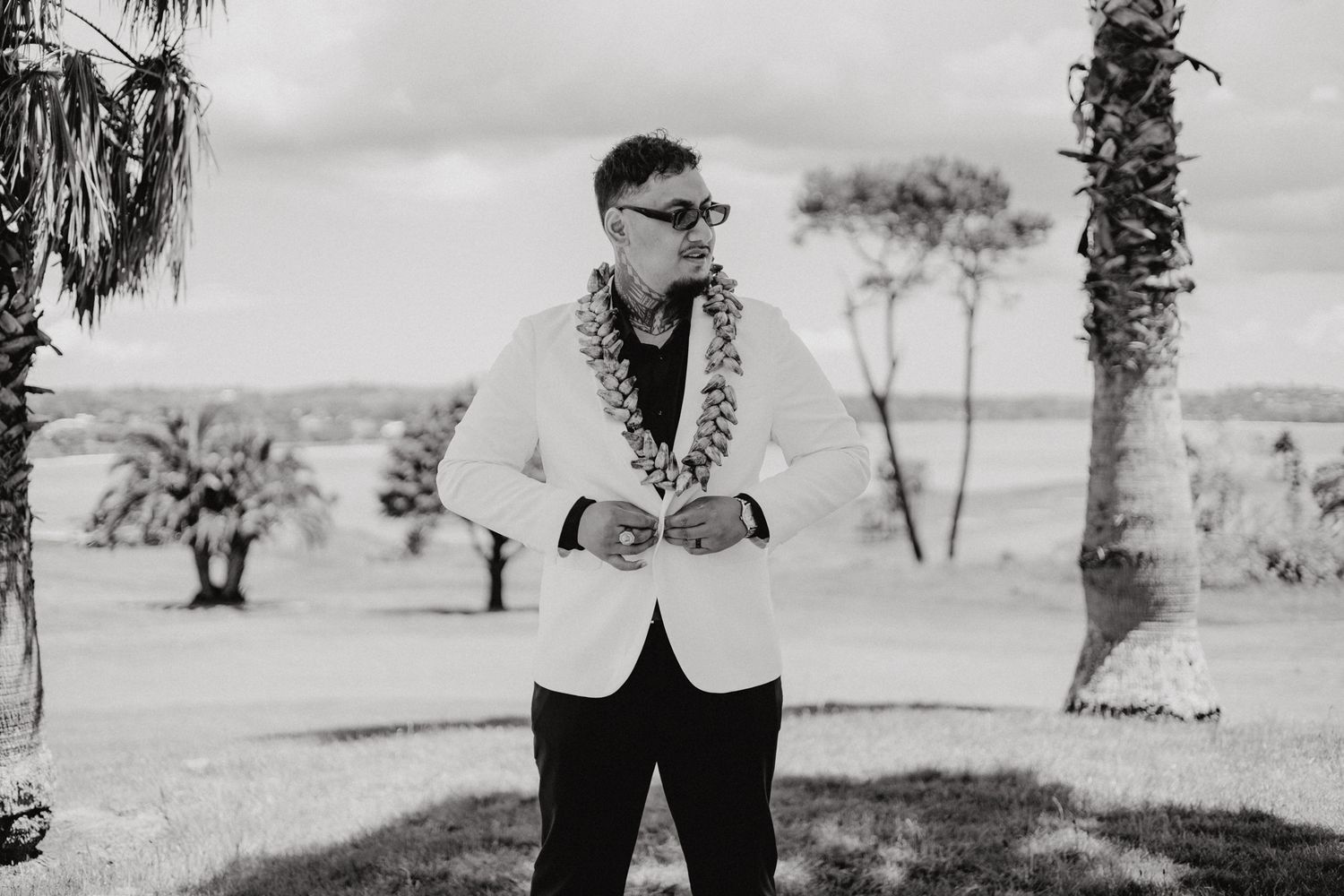 Person in white blazer and lei poses in black and white photo with palm trees in background.
