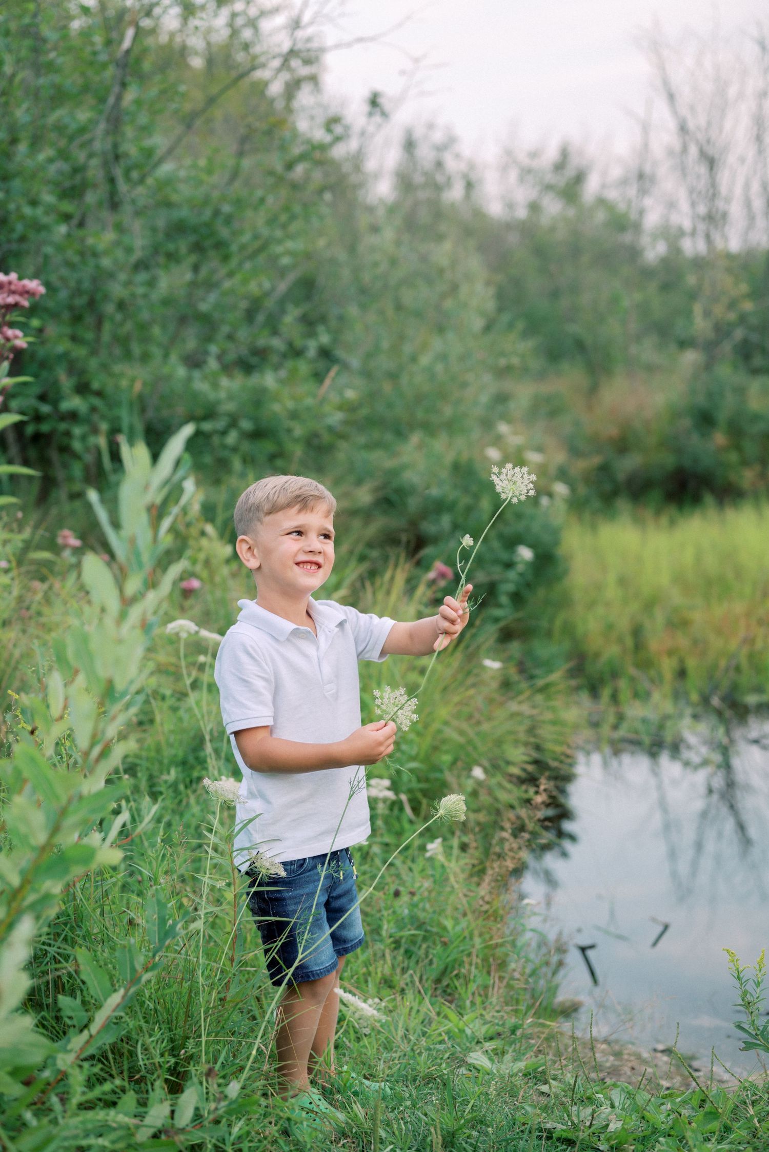 Young child exploring nature while holding wildflowers by a tranquil pond on a summer day.