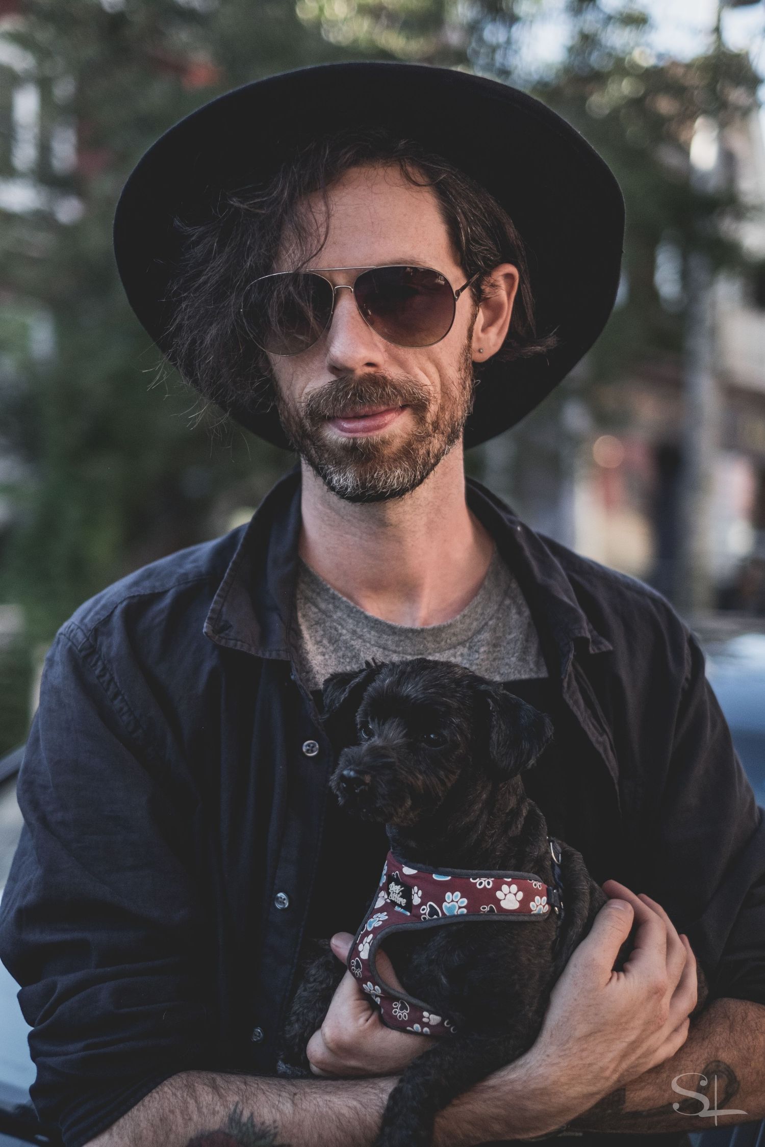 a man with a wide brim hat, dark hair and sunglasses holding a black puppy