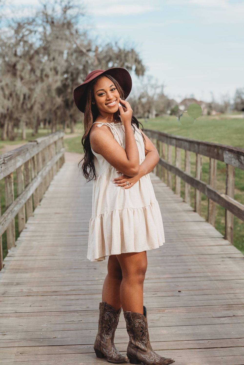High School Senior Girl wearing a Burgundy Hat, Beige Dress, and Cowboy boots on bridge in Sienna Plantation