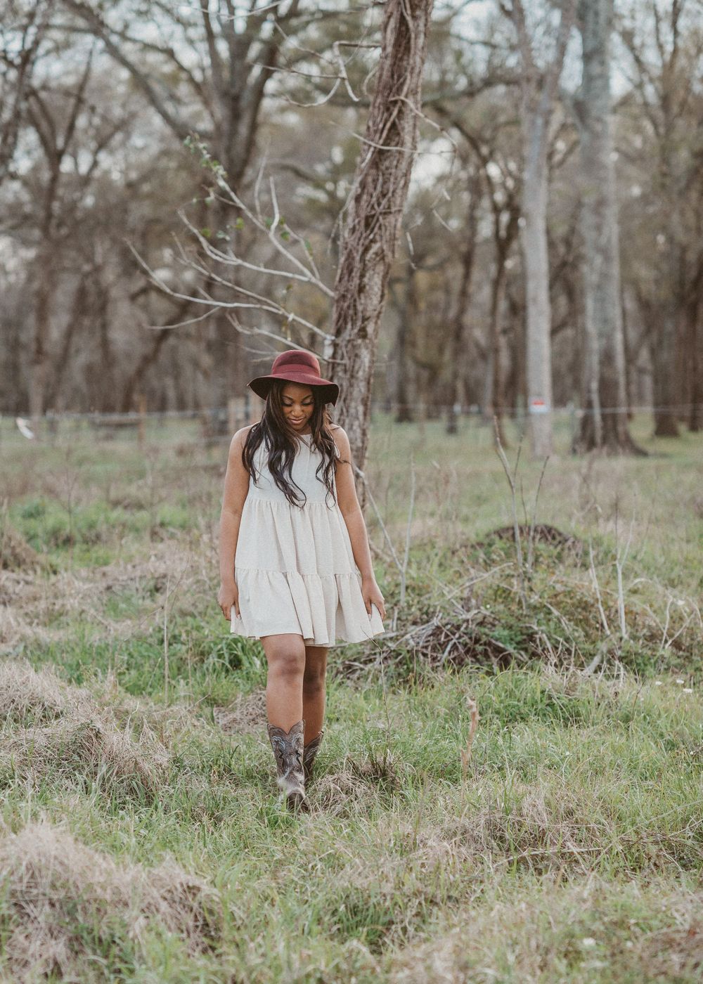 RidgePoint High School Senior Girl in a Burgundy Hat, Beige Dress, and Cowboy boots in woods in Sienna Plantation