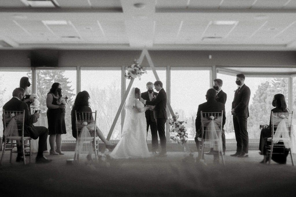 A wedding ceremony takes place in a bright modern room with large windows and guests seated on chairs.