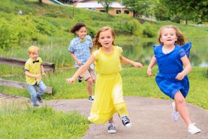 Children in colorful dresses running and playing on grassy hill near pond.