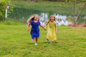 Two children in blue and yellow dresses running together on grass near water.