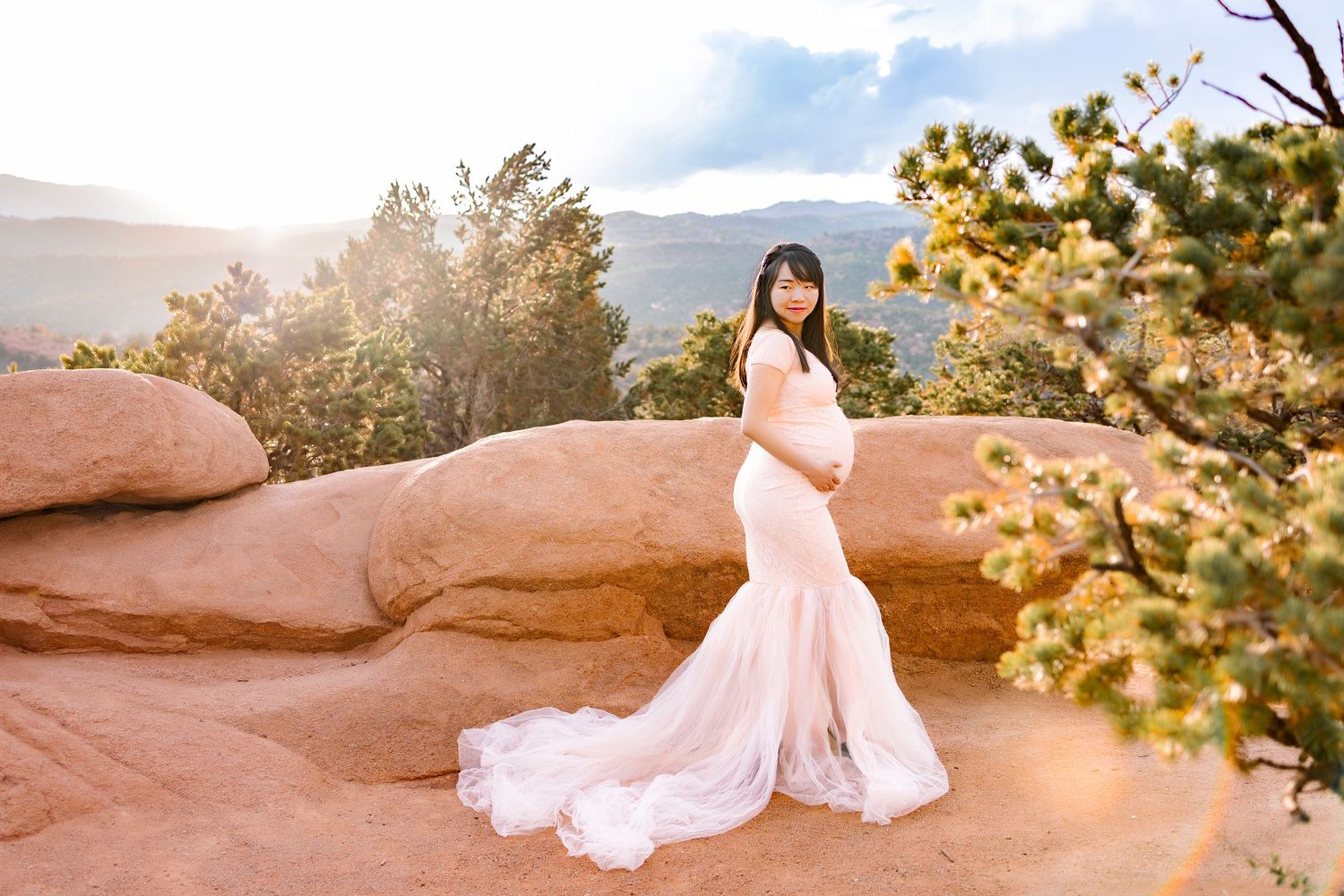 Pregnant woman in long pink dress, she is holding the bottom of her bump, looking down over right shoulder, side stance