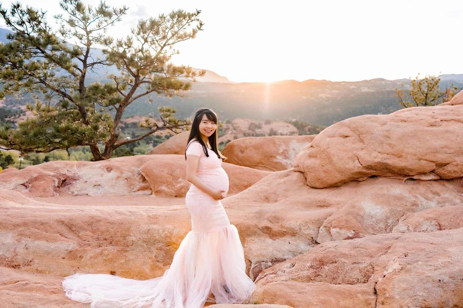 Pregnant woman, side stance, holding bottom of bump, looking at camera smiling, tree behind left, sunset, stood on red