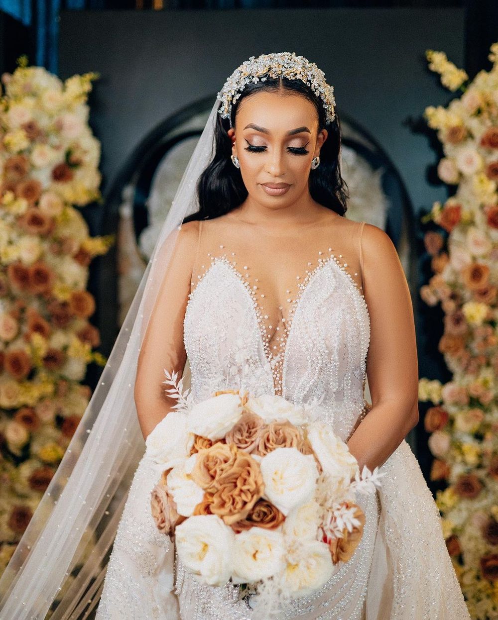 A bride in an elegant lace wedding gown with crystal headpiece holds a bouquet of cream and blush roses against floral decor.