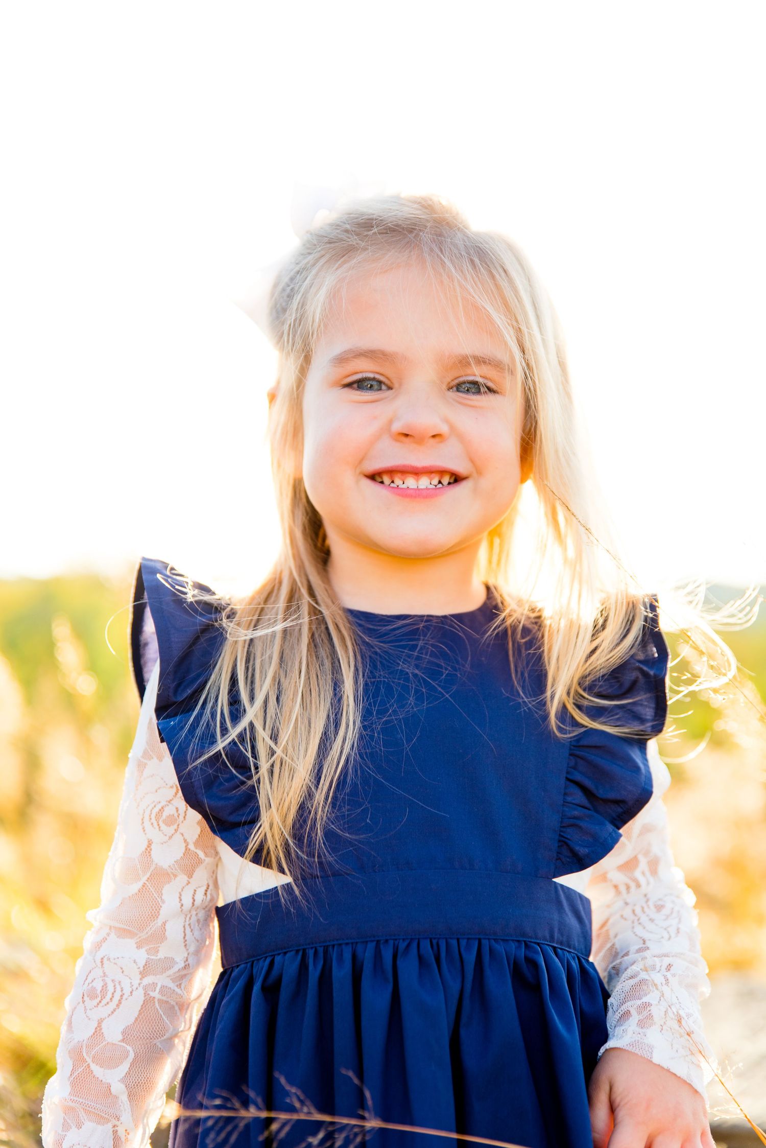 young blonde girl in navy dress and white lace long sleeve shirt smiling in front of the sunset