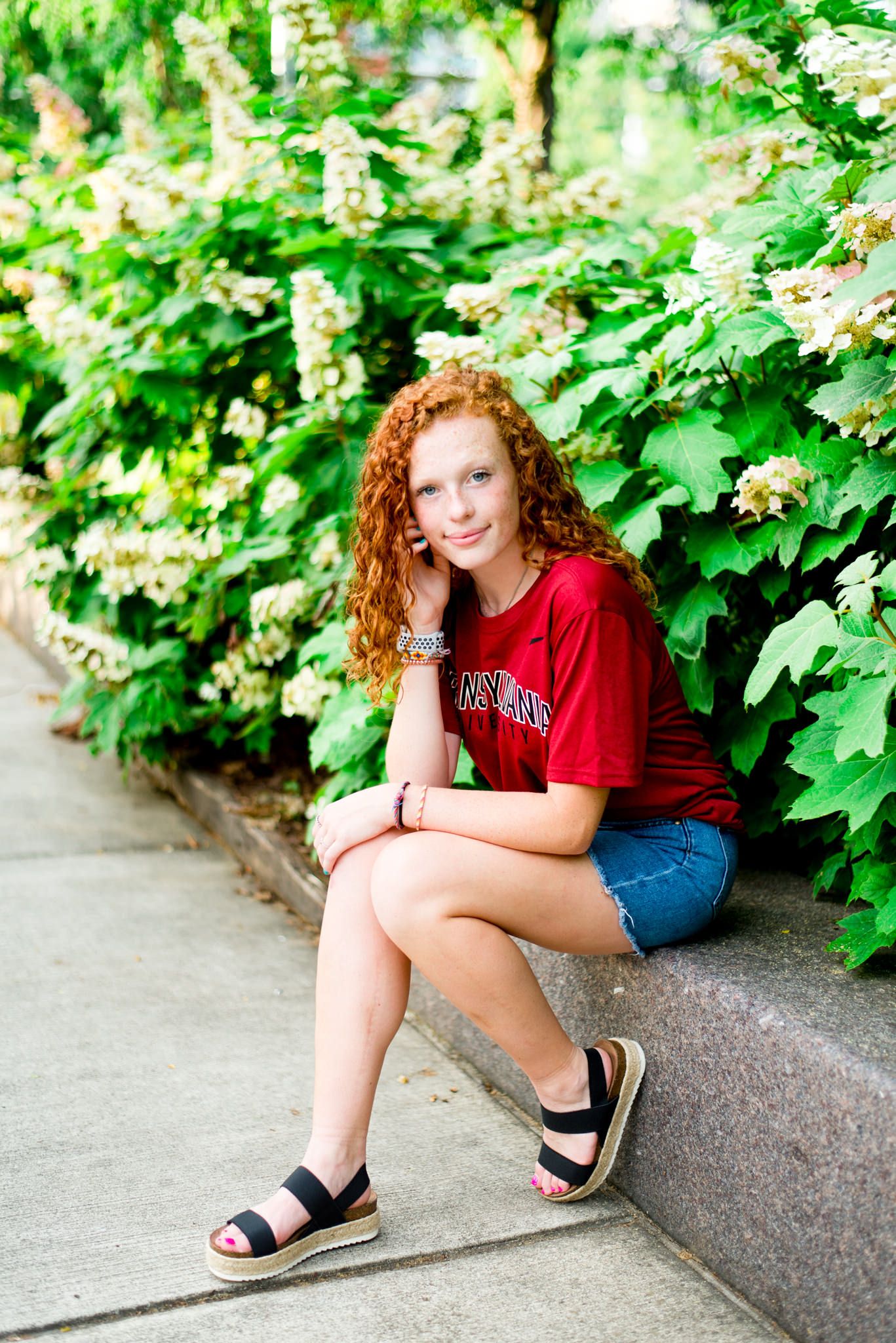 young woman with curly red hair in a red Transylvania shirt and jean skirt sitting in front of hydrangeas at Smale Park