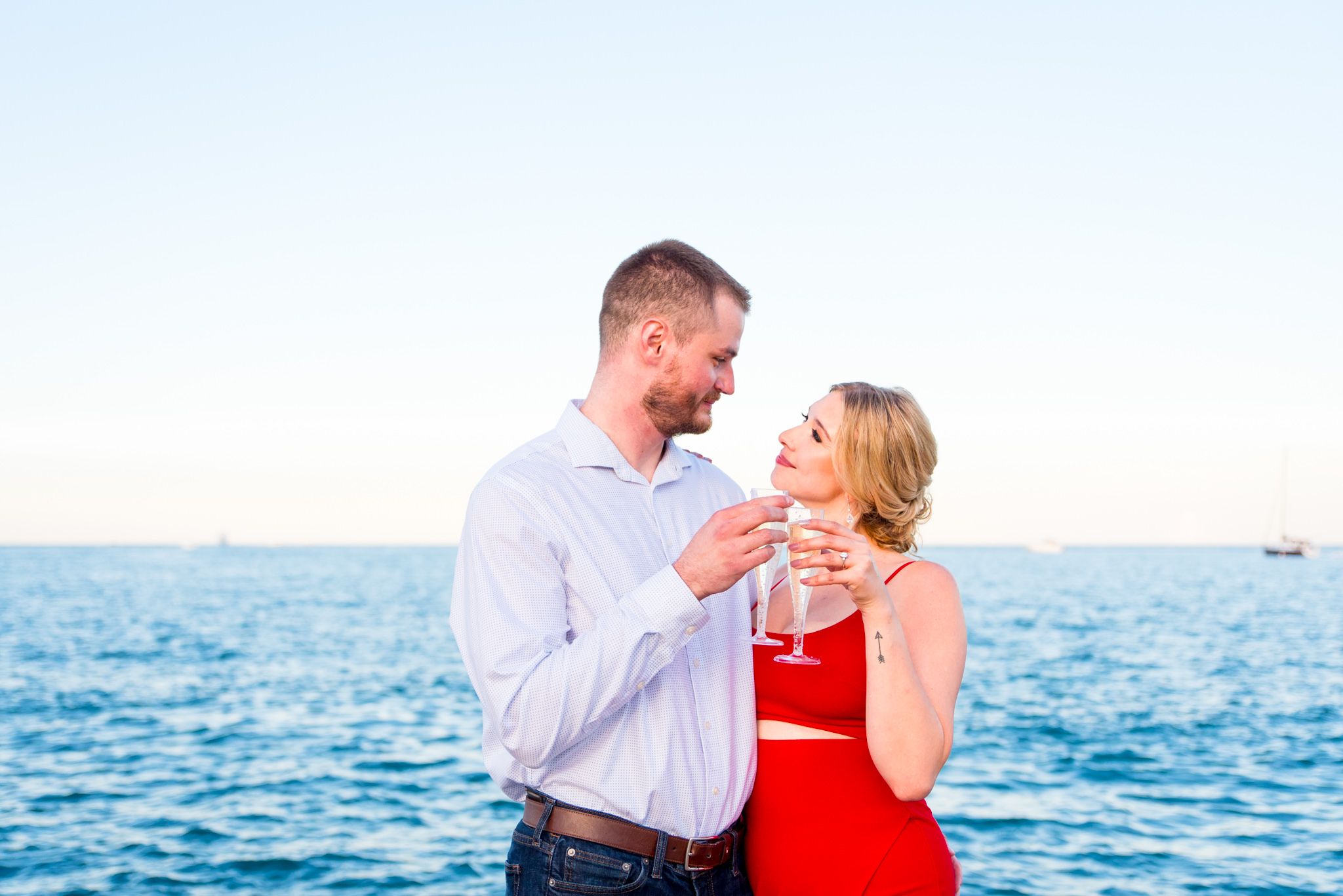 engaged couple in red dress and white shirt smiling at each other with champagne at sunset in Chicago engagement picture