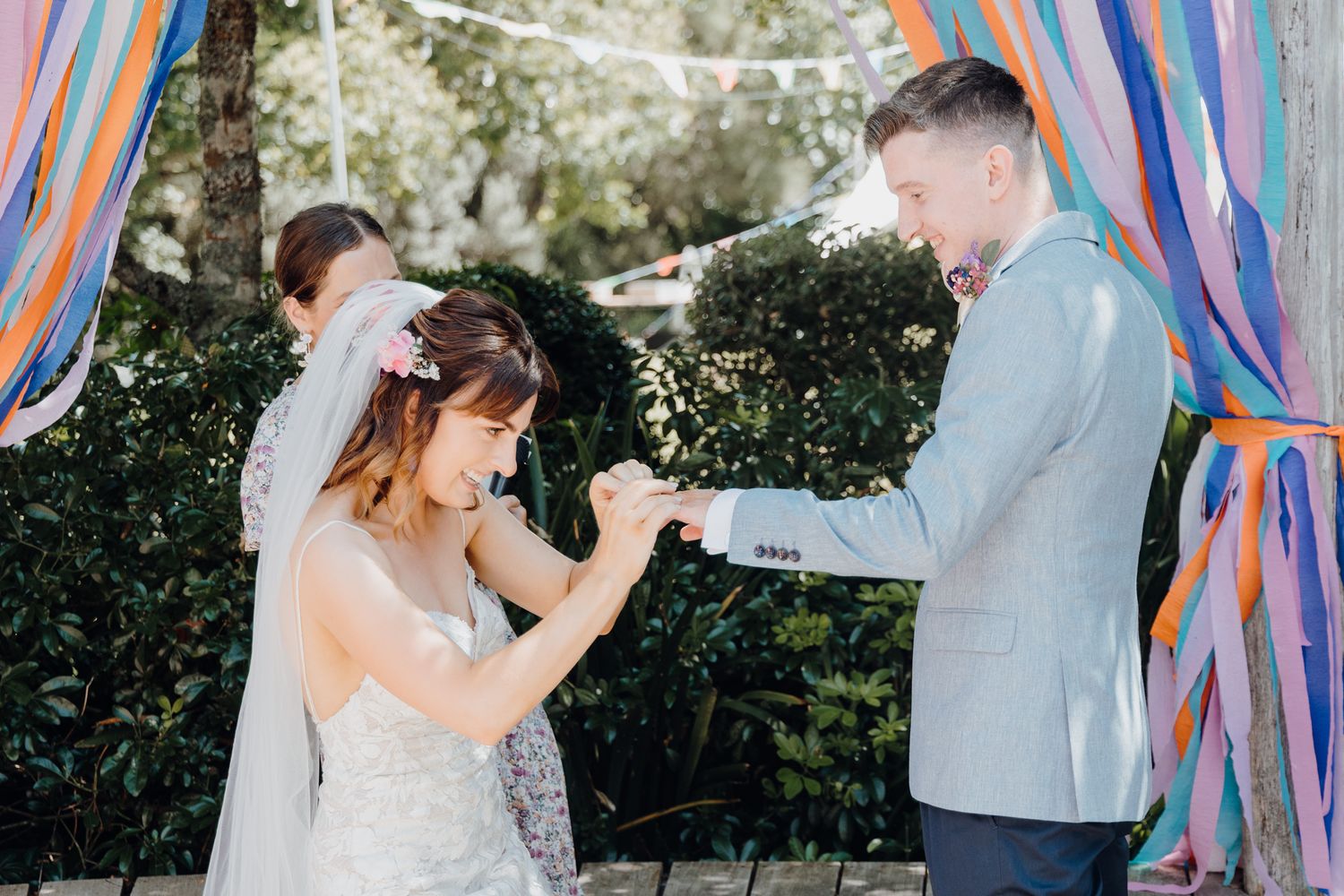 A bride in white lace reads her vows during an outdoor ceremony with colorful ribbon decorations.