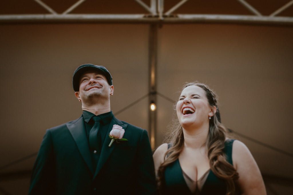 Wedding guests laugh and dance together under exposed wooden beams at an indoor reception.