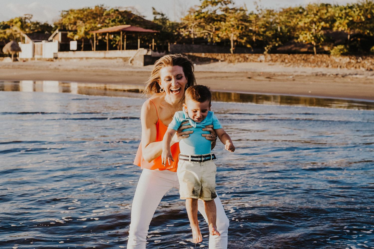 mother holds son over the water on the beach while laughing
