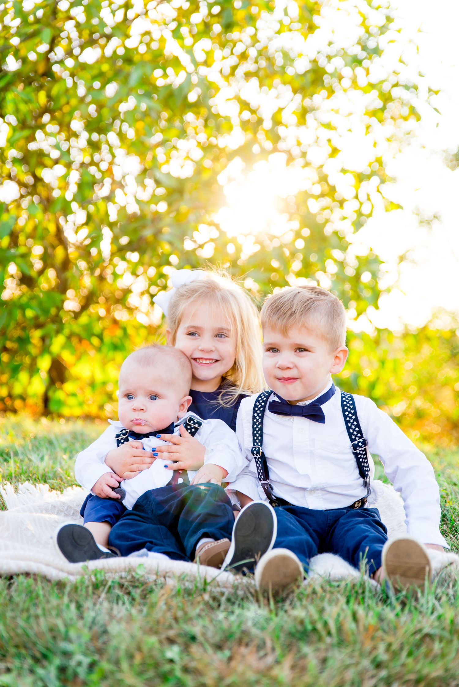 two young children and a baby boy wearing navy and white smiling in front of a blurry green bush at sunset in Chicago