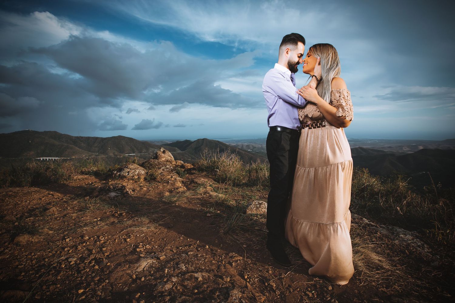 A romantic couple embraces on a mountaintop against a dramatic cloudy sky during golden hour.