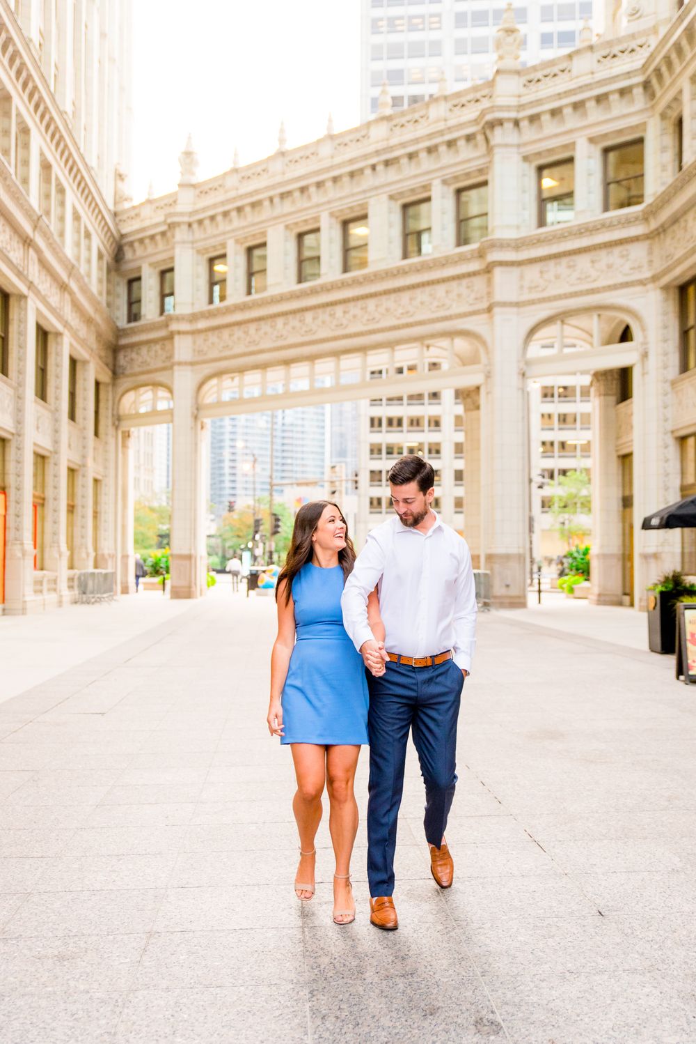 woman with brown hair in a light blue dress holding hands with fiance in front of the wrigley building in chicago
