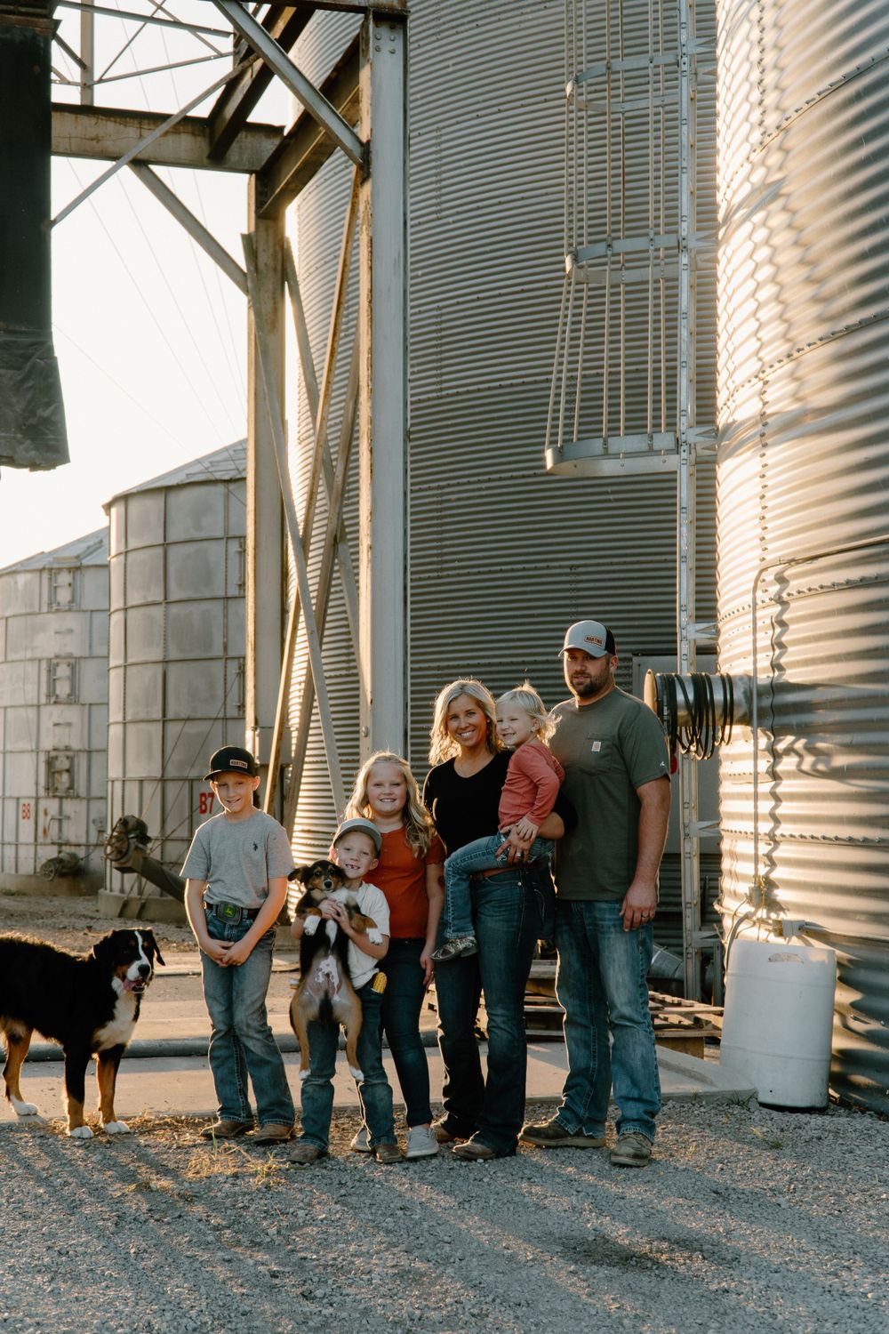 Family gathering with their dogs near metal grain storage silos at dusk.