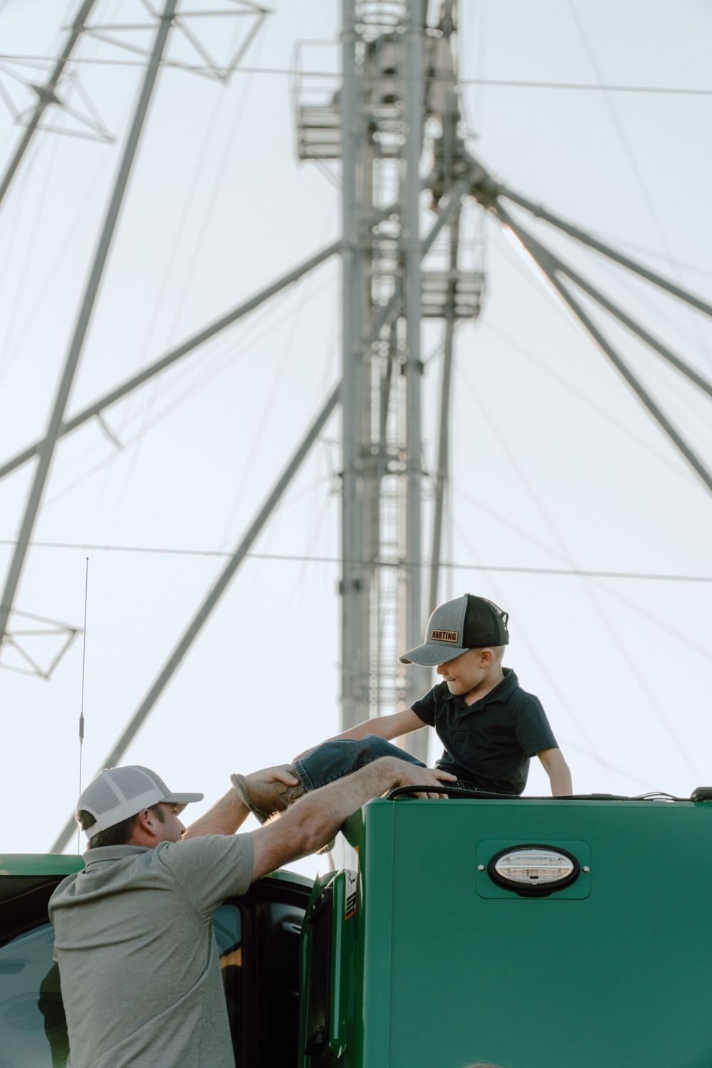 Adult helping child climb onto large green farming combine harvester in an agricultural setting.