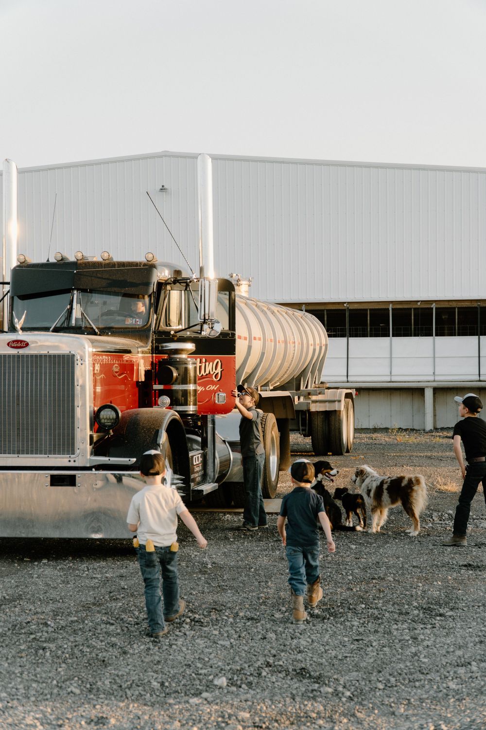 Children approaching a red semi truck with a tanker trailer while a dog watches nearby.