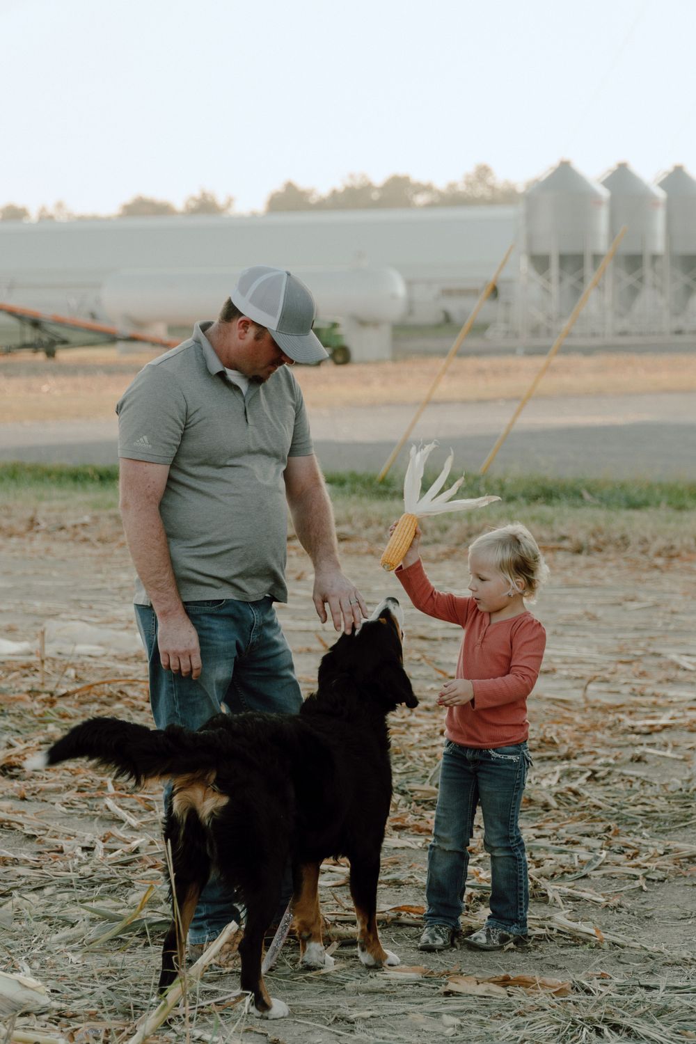 Small child and adult interact with a black dog near grain storage silos on a farm.